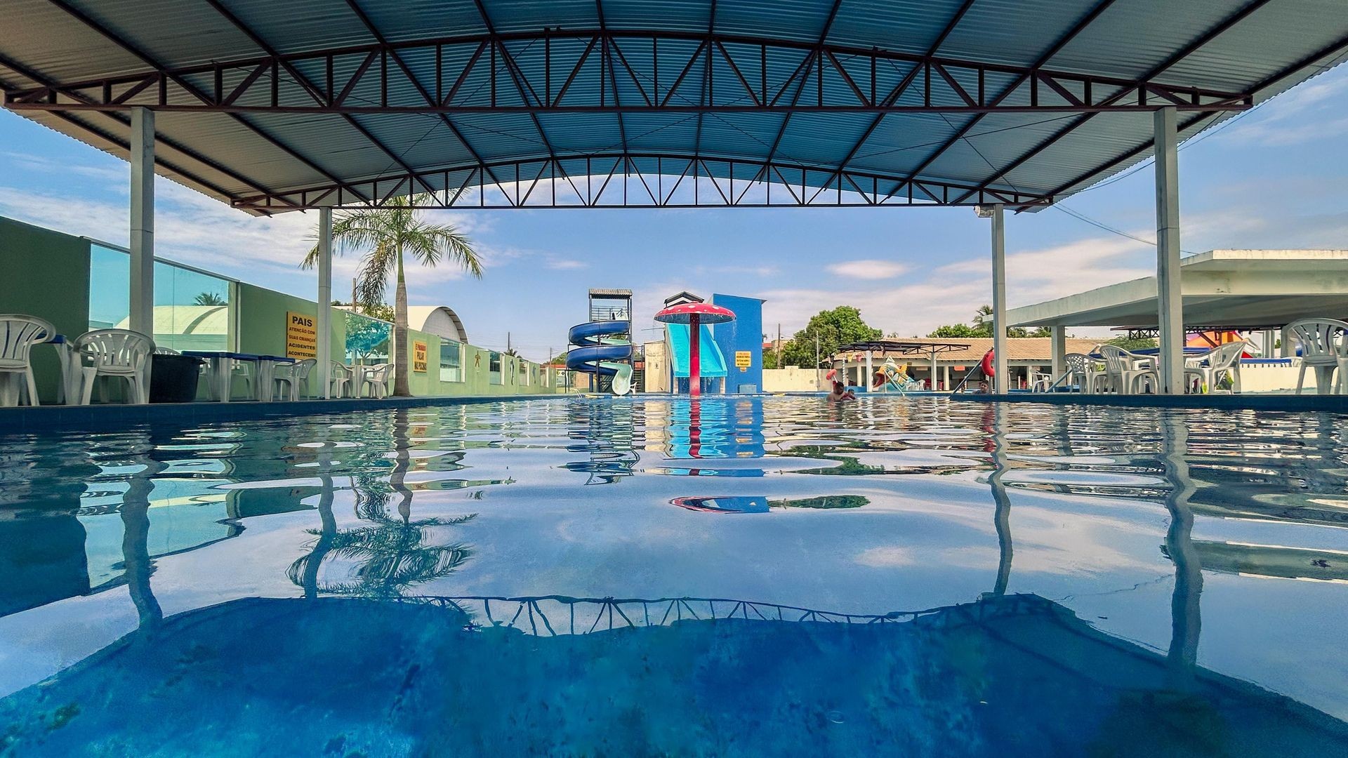 Imagem de uma piscina grande e cristalina no Balneário Central Park. A água reflete o céu azul e as estruturas do clube ao redor. Ao fundo, há toboáguas e cadeiras organizadas ao longo da piscina. O ambiente transmite tranquilidade e diversão, ideal para relaxar e se refrescar.