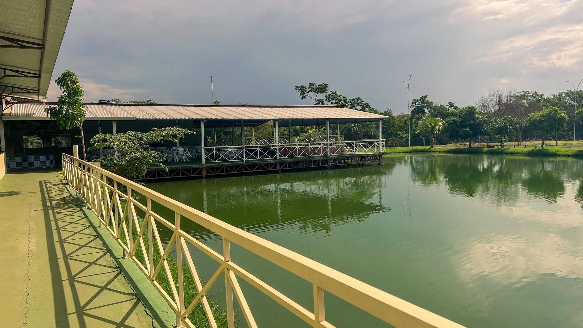 Vista de uma passarela com guarda-corpo ao lado de um lago verde, cercado por vegetação. Ao fundo, há um grande salão com telhado e mesas, parte da estrutura do Balneário Central Park, proporcionando um ambiente tranquilo e integrado à natureza.